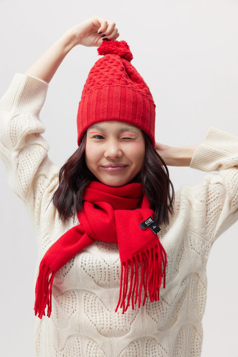 A woman wearing a bright red knit beanie with a pom-pom and a matching red scarf, posed against a light gray studio background. The scarf has a small black brand tag reading 'GEE'.