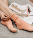 Model wearing ribbed peach cashmere socks, adjusting the folded cuff beside stacked books