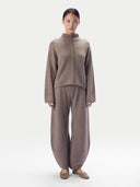 A model stands against a light gray studio background wearing a matching taupe-brown knit two-piece set consisting of a mock-neck long-sleeve top with a central braided detail and wide-leg pants.