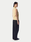 A model shown from the side profile wearing a pale yellow sleeveless mock-neck knit sweater vest and dark charcoal wide-leg trousers against a light gray studio background.