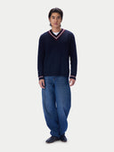 A man wearing a navy blue cable-knit V-neck sweater with red and white trim, paired with loose-fit blue jeans and dark shoes, standing against a light gray studio background.