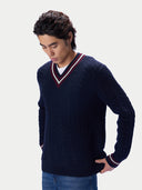 A man wearing a navy blue cable-knit V-neck sweater with red and white trim, layered over a white undershirt and paired with blue jeans, set against a plain light gray background.