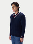 A man wearing a navy blue cable-knit V-neck sweater with red and white striped trim, paired with blue jeans, photographed against a light gray studio background.