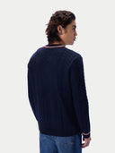 A man shown from the back wearing a navy blue cable-knit sweater with red and white striped trim at the collar and cuffs, paired with blue jeans, against a light gray studio background.