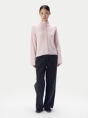 A model stands against a light gray studio background wearing a pale pink mock-neck knit top with a decorative braided front detail and black wide-leg trousers.