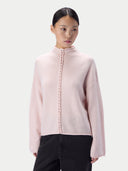 A model wearing a pale pink knit mock-neck sweater with a decorative braided front detail, paired with black pants, against a light gray studio background.