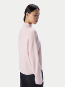 A woman shown in side profile wearing a pale pink mock-neck knit sweater and black pants against a clean light gray studio background.