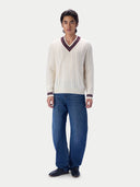 A man modeling a cream cable-knit V-neck sweater with dark red and navy striped trim, paired with blue jeans and black shoes, against a light gray studio background.