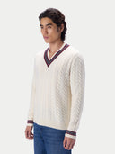 A man wearing a cream cable-knit V-neck sweater with navy and red striped trim, paired with blue jeans, photographed against a light gray studio background.