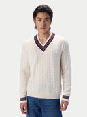 A man wearing a cream cable-knit V-neck sweater with navy and red striped trim, paired with blue jeans, against a light gray studio background.