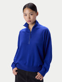 A woman modeling a bright royal blue quarter-zip knit sweater with a stand collar, paired with black pants, against a light gray studio background.