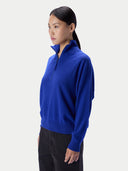 A woman wearing a bright royal blue quarter-zip mock-neck sweater and black pants, shown in profile against a light gray studio background.
