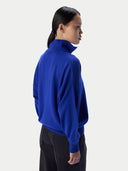 A woman shown from the back and side wearing a bright royal blue mock-neck sweater and black pants against a light gray studio background.