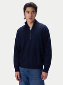A man wearing a navy blue quarter-zip knit sweater and blue jeans, standing against a light gray studio background.