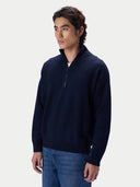 A man wearing a navy blue quarter-zip knit sweater and blue jeans, standing against a light gray studio background.