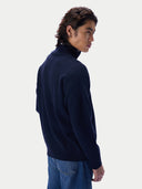 A man wearing a dark navy blue turtleneck sweater and blue jeans, shown from a side-back angle against a light gray studio background.