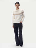 A model stands against a light gray studio background wearing a cream knit sweater with a brown Fair Isle pattern across the chest and shoulders, paired with dark charcoal wide-leg pants and black shoes.
