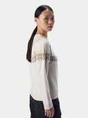 A woman wearing a cream-colored knit sweater with a brown geometric Fair Isle pattern across the shoulders and upper chest, paired with black pants, photographed against a light gray studio background.