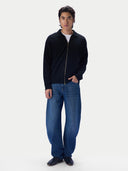 A man modeling a black zip-up jacket with a gold zipper, worn over a white t-shirt and paired with blue jeans and black shoes, against a plain light gray background.