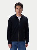 A man wearing a black zip-up knit polo cardigan over a white t-shirt and blue jeans, standing against a light gray studio background.