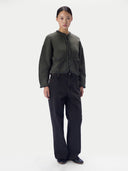 A model stands against a plain light gray studio background wearing a dark olive green cropped jacket with black chain trim and black wide-leg jeans.