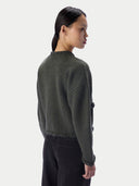 A woman shown from the back wearing a dark olive green cropped knit sweater with braided trim at the collar and hem, paired with black pants, against a light gray studio background.