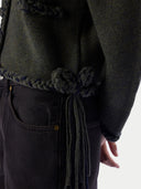 Close-up of a person wearing a dark olive green knit sweater with a thick braided cord tied around the waist, paired with dark jeans against a plain light background.