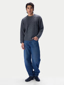 A man modeling a dark gray cable-knit crewneck sweater paired with loose blue jeans and black shoes, standing against a plain light gray studio background.