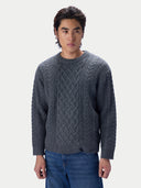 A man wearing a dark gray cable-knit crewneck sweater and blue jeans, standing against a light gray studio background.