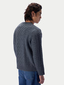 A man wearing a dark gray cable-knit sweater and blue jeans, shown from a side-back angle against a light gray studio background.
