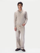 A man modeling a beige ribbed knit V-neck sweater with contrasting dark trim, paired with beige trousers and dark brown shoes, against a plain light gray studio background.