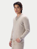 A man wearing a ribbed knit beige sweater with a V-neck and striped cuffs, paired with beige pants, photographed against a light gray studio background.
