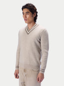 A man wearing a beige ribbed knit V-neck sweater with striped trim on the collar and cuffs, paired with light beige trousers, photographed against a light gray studio background.