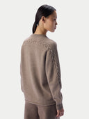 A model shown from the back wearing a taupe knit sweater with braided cable detailing along the shoulders and sleeves, paired with matching knit pants, against a light gray studio background.