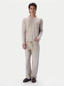 A man modeling a light beige knit crewneck sweater and matching beige trousers with dark brown suede shoes, set against a plain light gray studio background.