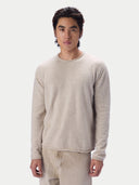 A man wearing a light beige crewneck knit sweater and matching beige trousers, standing against a solid off-white background.