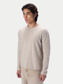 A man wearing a light beige crewneck knit sweater and matching beige trousers, photographed against a clean light gray studio background.