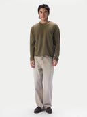 A man modeling a long-sleeve crewneck sweater in olive green, paired with light beige trousers and dark brown shoes, against a plain white studio background.