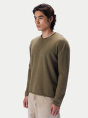 A man wearing a muted olive green long-sleeve knit sweater and light beige trousers, standing against a plain light gray studio background.