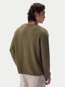 A man shown from the back wearing a muted olive green knit sweater and light beige trousers, photographed against a clean off-white studio background.
