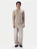 A man wearing a light beige knit mock-neck sweater and matching beige trousers, standing against a plain light gray studio background.