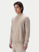 A man wearing a light beige knit mock-neck sweater and matching trousers, photographed against a clean light gray studio background.