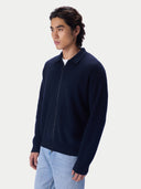 A man wearing a dark navy blue zip-up knit polo cardigan over a white t-shirt and light blue jeans, photographed against a clean, light gray studio background.
