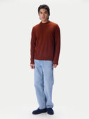 A man wearing a rust-brown knit mock-neck sweater, light blue jeans, and dark navy loafers, standing against a plain white studio background.