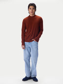 A man wearing a rust-brown mock neck knit sweater, light blue jeans, and navy blue loafers, standing against a solid off-white background.