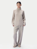 A model wearing a light beige knit turtleneck sweater and matching wide-leg trousers, standing against a solid light gray background.