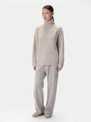 A model wearing a light beige ribbed turtleneck sweater and matching wide-leg trousers, standing against a plain light gray studio background.