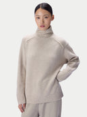 A model wearing a light beige knit turtleneck sweater and matching trousers, photographed against a clean light gray studio background.