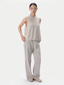 A model stands against a light gray studio background wearing a sleeveless, mock-neck knit top and matching wide-leg trousers in a light beige color, paired with beige slip-on shoes.