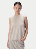 A model wearing a light beige sleeveless knit top with a mock neck and light beige trousers, photographed against a solid light gray background.